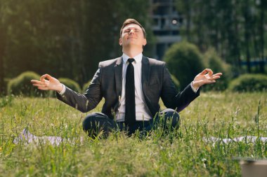 Genç adam kibarca çimlerin üzerinde oturuyor, uzaktan çalışıyor. Küçük işletme konsepti, namaste yapan serbest çalışan bir adam, öğle yemeği sırasında ucuz bir huzur.