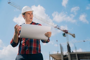 Beyaz koruyucu kasklı bir adam portresi. Elinde bir bina inşaatı kağıdıyla. Doğanın mavi gökyüzüne sahip bir vincin arka planındaki akıllı mühendis kavramı..