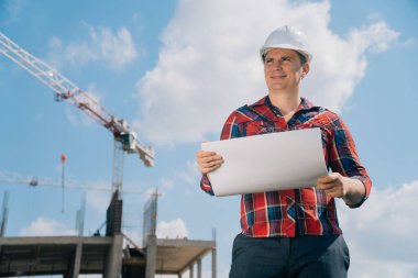 Beyaz koruyucu kasklı neşeli bir adamın portresi. Elinde bir binanın arka planında kağıt var. Mavi gökyüzü olan bir vincin arka planında akıllı bir mühendis kavramı.