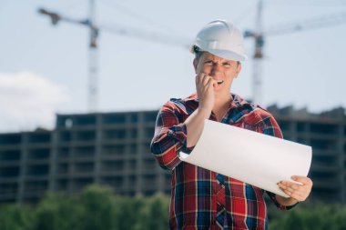 Beyaz koruyucu kasklı bir adam portresi. Elinde bir bina inşaatı kağıdıyla. Doğanın mavi gökyüzüne sahip bir vincin arka planındaki akıllı mühendis kavramı..