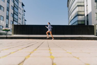 Spor kıyafetleri içinde şık bir genç kız, açık havada gökdelenler arasında koşup kilo vermeye çalışıyor. Yaz günbatımı spor konsepti, kilo verme sporları