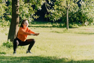 Spor giyimli bir kız dışarıda çömeliyor, kilo vermek için kalça kaslarını çalıştırıyor. Yoga, gün batımında paspas kullanarak fonksiyonel eğitim kavramı.