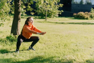 Spor giyimli bir kız dışarıda çömeliyor, kilo vermek için kalça kaslarını çalıştırıyor. Yoga, gün batımında paspas kullanarak fonksiyonel eğitim kavramı.