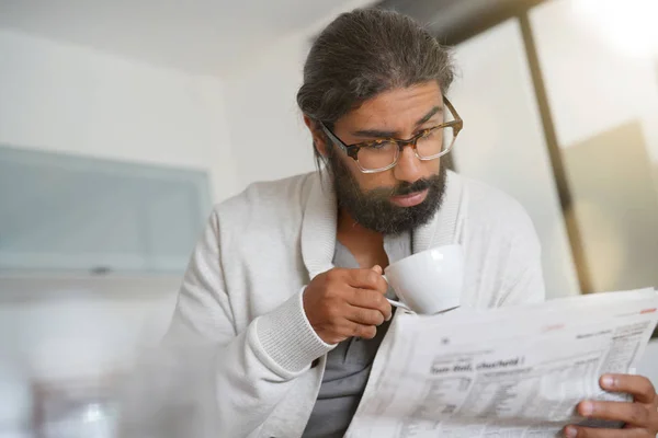 Dark-haired bearded man at home reading newspaper