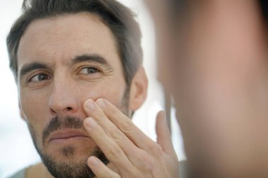 Handsome man applying face cream looking in mirror clipart
