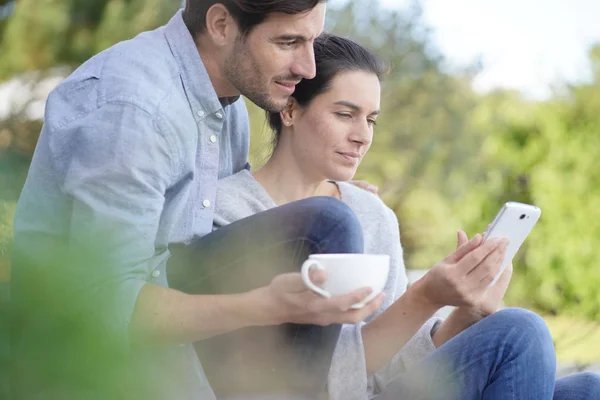 放松的年轻夫妇坐在外面 拿着杯子和手机 — 图库照片