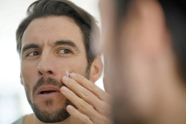Bonito Homem Aplicando Creme Facial Olhando Espelho — Fotografia de Stock
