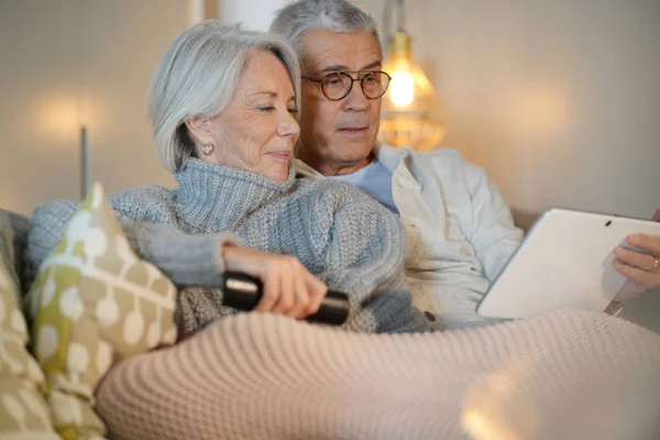 在家里的老年夫妇在沙发上放松看电视和看平板电脑 — 图库照片