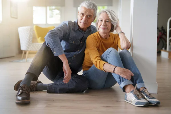 Relaxed senior couple sitting on floor in modern home looking at camera