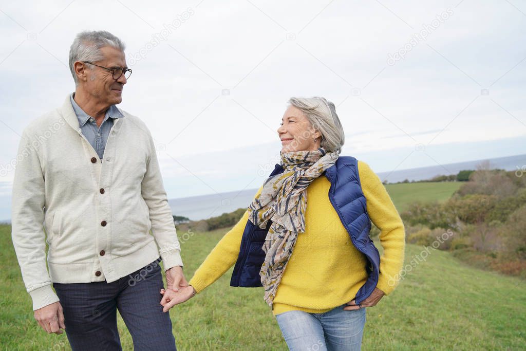  Healthy senior woman holding husband's hand and leading way on countryside walk                          