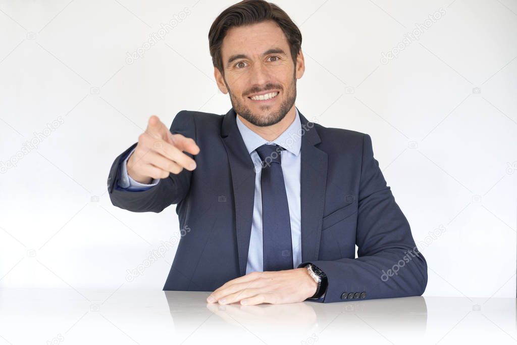 Isolated attractive businessman sitting at desk in suit pointing at camera