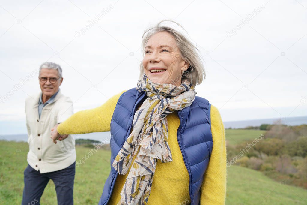  Healthy senior woman holding husband's hand and leading way on countryside walk                          