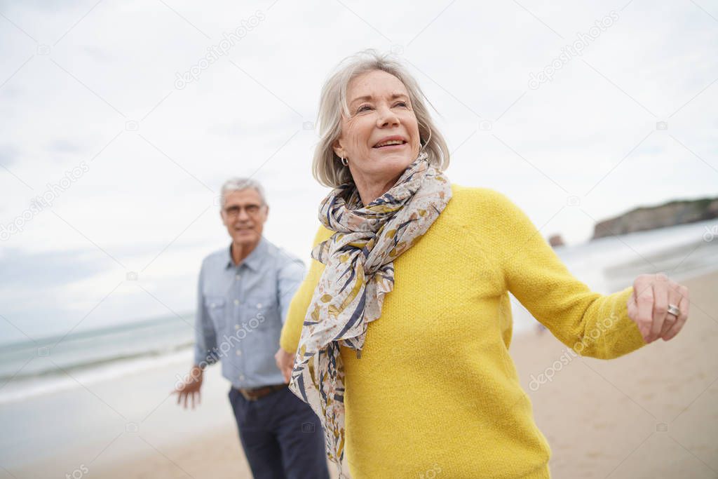 Vibrant senior woman holding husband's hand and leading the way on beach walk                             
