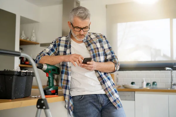 中年の男性がモダンなキッチンの棚を固定しながら 携帯電話をチェック — ストック写真