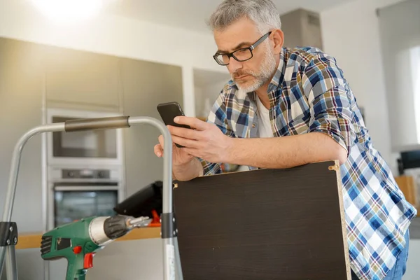 Mature man checking phone whilst fixing shelf in modern kitchen