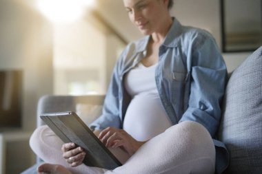 Dijital tablet kullanan hamile kadın, kanepede dinleniyor