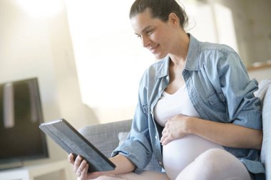 Dijital tablet kullanan hamile kadın, kanepede dinleniyor