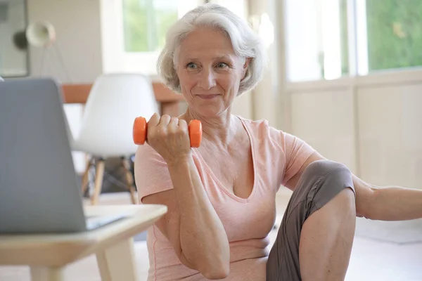 Senior woman doing fitness exercises with virtual class, holding dumbbells