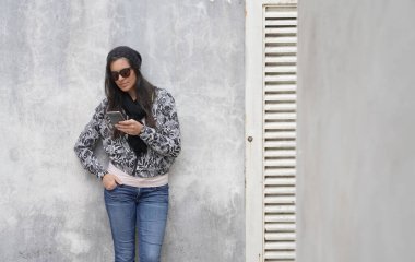 Siyah güneş gözlüğü takan trend bir kadın beton duvarda duruyor ve akıllı telefon kullanıyor.