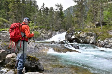 Kırmızı sırt çantalı ve kırmızı ceketli uçan balıkçı yazın yüksek bir dağ nehrinde balık tutuyor.