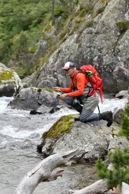 Uçan balıkçı alabalığı, yaz aylarında yüksek dağlarda yürüyüş çantası ve turuncu bir ceketle balık tutuyor.