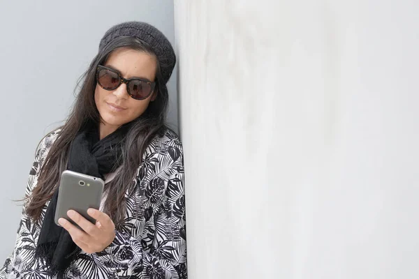 trendy woman wearing black sunglasses standing on concrete wall and using smartphone