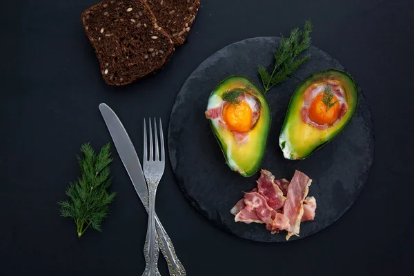 two baked avocado halves with egg on black stone Board