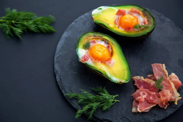 Tasty baked avocado halves with egg and sliced becon on black stone Board,close up