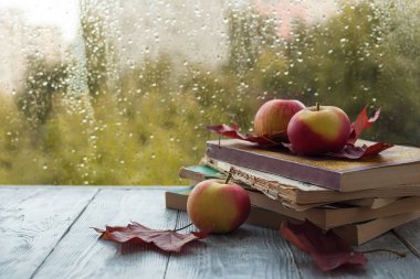 ıslak bir pencere yakınında ahşap gri bir arka plan üzerinde akçaağaç yaprakları ile eski kitap ve elma yığını. Sonbahar natürmort.