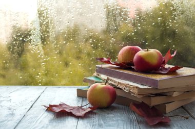 ıslak bir pencere yakınında ahşap gri bir arka plan üzerinde akçaağaç yaprakları ile eski kitap ve elma yığını. Sonbahar natürmort.