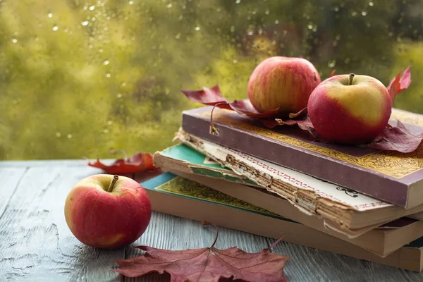 ıslak bir pencere yakınında ahşap gri bir arka plan üzerinde akçaağaç yaprakları ile eski kitap ve elma yığını. Sonbahar natürmort.