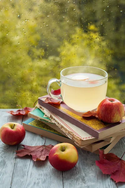 ıslak bir pencere yakınında ahşap bir masa üzerinde bir elma ve akçaağaç yaprakları ile eski kitap ve içki yığını.