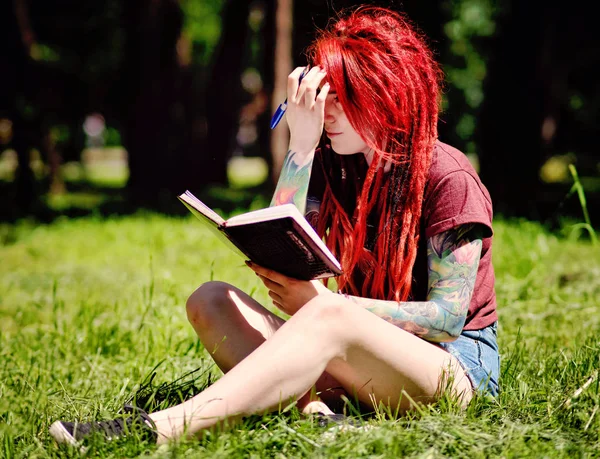 Young female student with red hair and dreadlocks studying in a summer park with a notebook and pen
