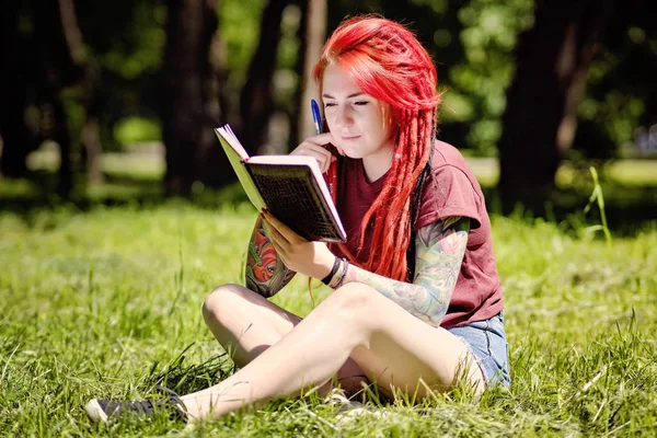 Young female student with red hair and dreadlocks studying in a summer park with a notebook and pen