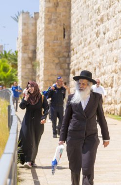 Jerusalem, Israel - June 14, 2018: Old Haridi Jewish man and young Muslim girl walking around the old city of Jerusalem. Jerusalem hosts believers of the three biggest monotheistic religions. clipart