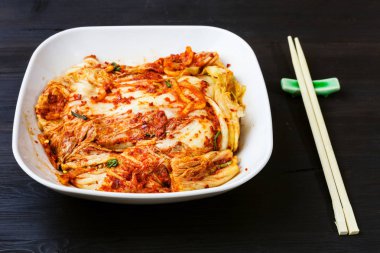 Kore mutfağı - Kimchi aperatifi (baharatlı nappa lahanası) beyaz kâse ve koyu kahverengi ahşap masa üzerinde çubuklar