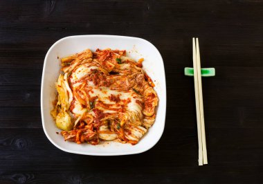 Kore mutfağı - Kimchi aperatiflerinin üst görüntüsü (baharatlı nappa lahanası) beyaz kâse ve koyu kahverengi ahşap masa üzerinde çubuklar