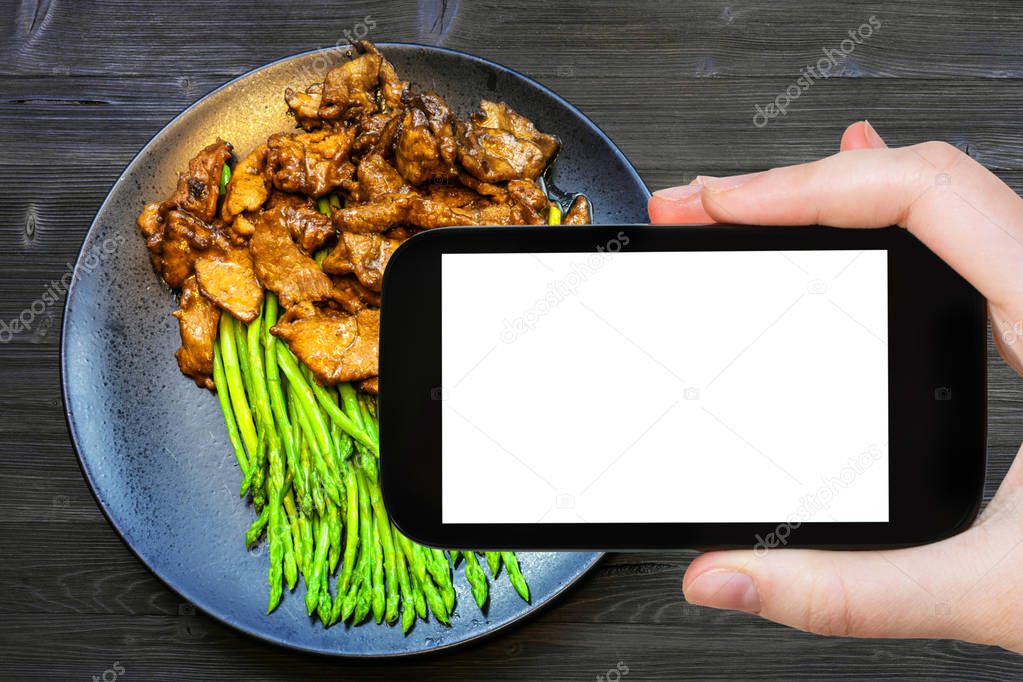 travel concept - tourist photographs of Chinese cuisine dish of Beef fried in soy sauce with green asparagus on smartphone with empty cutout screen with blank place for advertising