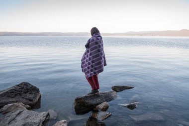 Ekoseli kız göl kıyısındaki taşın üzerinde duruyor.