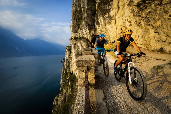 Cycling woman and man riding on bikes at sunrise mountains and Garda lake landscape. Couple cycling MTB enduro flow sentiero ponale trail track. Outdoor sport activity.