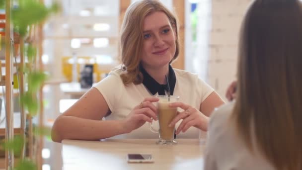 Dwie młode kobiety rozmawiają i piją kawę siedząc w kawiarni. — Wideo stockowe