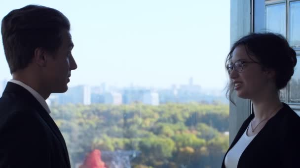 Два колеги стоячи біля вікна з великими міськими будівлями на задньому плані . — стокове відео