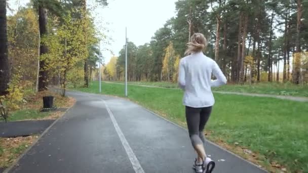 Вид ззаду на молоду блондинку, що біжить в осінньому парку . — стокове відео