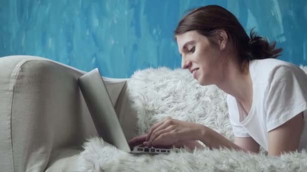Femme souriante utilisant un ordinateur portable à la maison tout en étant couché sur le canapé . — Video