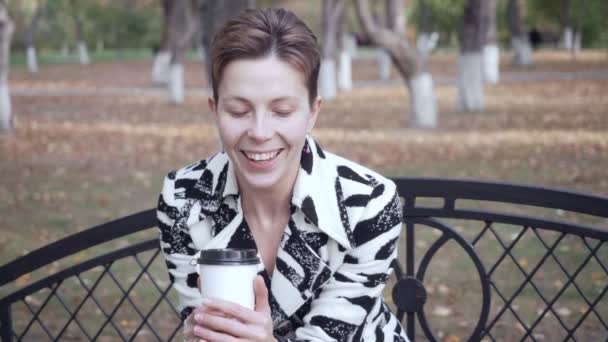 Mulher sentada em um banco em um parque, relaxando e bebendo café . — Vídeo de Stock