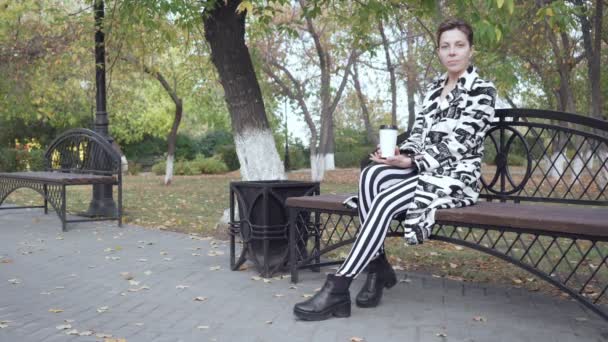 Kvinna sitter på en bänk i en Park, avkopplande och dricka kaffe. — Stockvideo