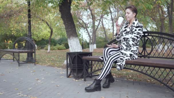 Kvinna sitter på en bänk i en Park, avkopplande och dricka kaffe. — Stockvideo