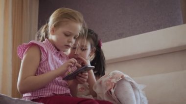 Yatak odasında yatakta oturan akıllı telefon kullanan iki küçük kız.