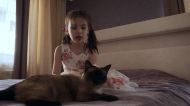 Yatakta oturan kedi okşayarak elbiseli küçük kız.