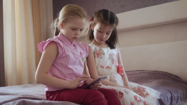 Δύο μικρά κορίτσια που χρησιμοποιούν smartphone κάθονται στο κρεβάτι στο υπνοδωμάτιο. — Αρχείο Βίντεο